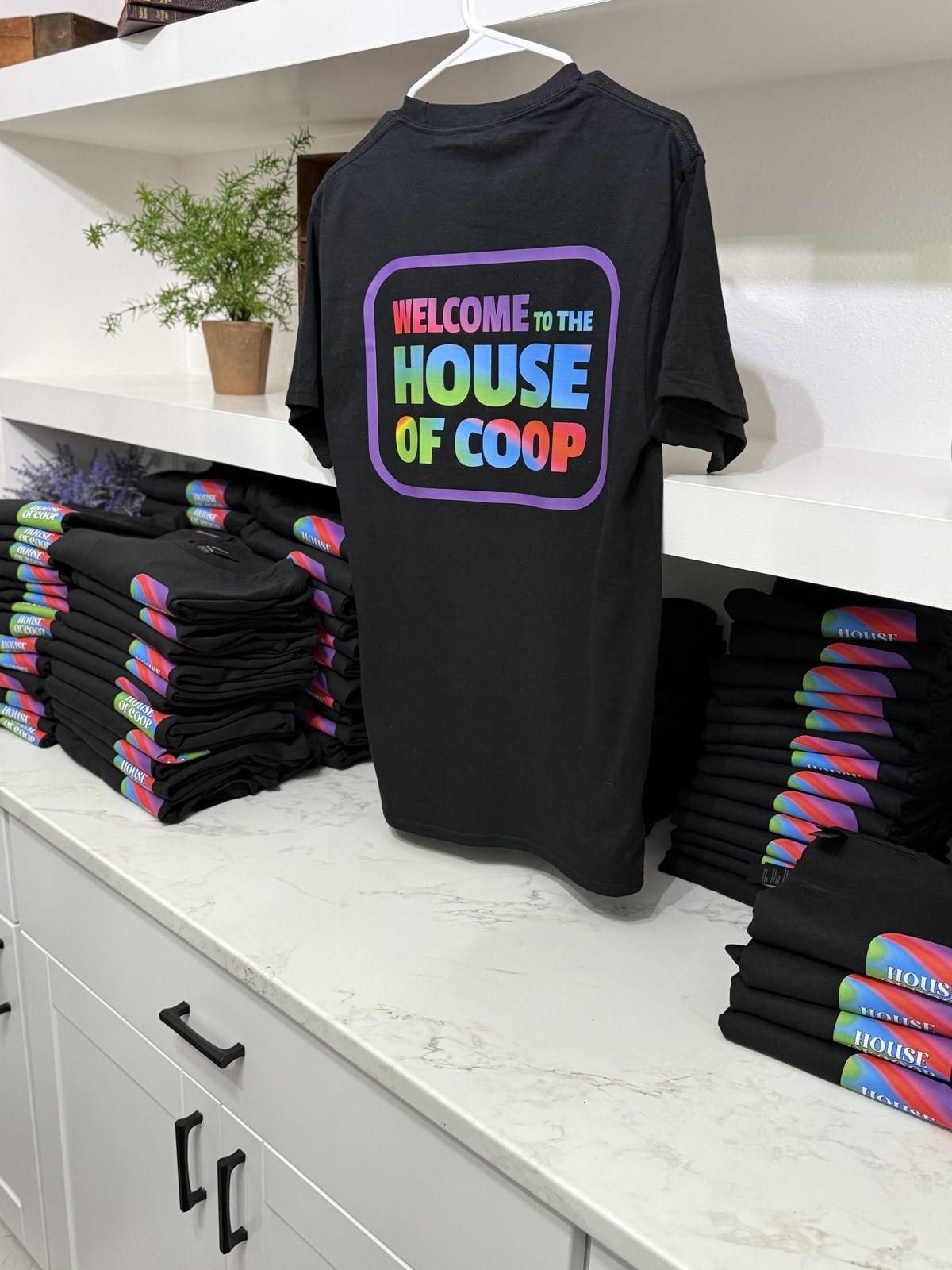 New black House of Coop shirts with colorful lettering, displayed beside stacks of folded shirts
