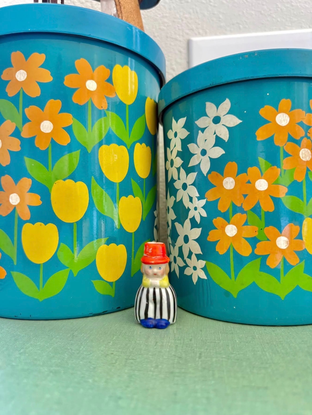 Tiny ceramic girl with striped pants and a red hat beside turquoise floral canisters