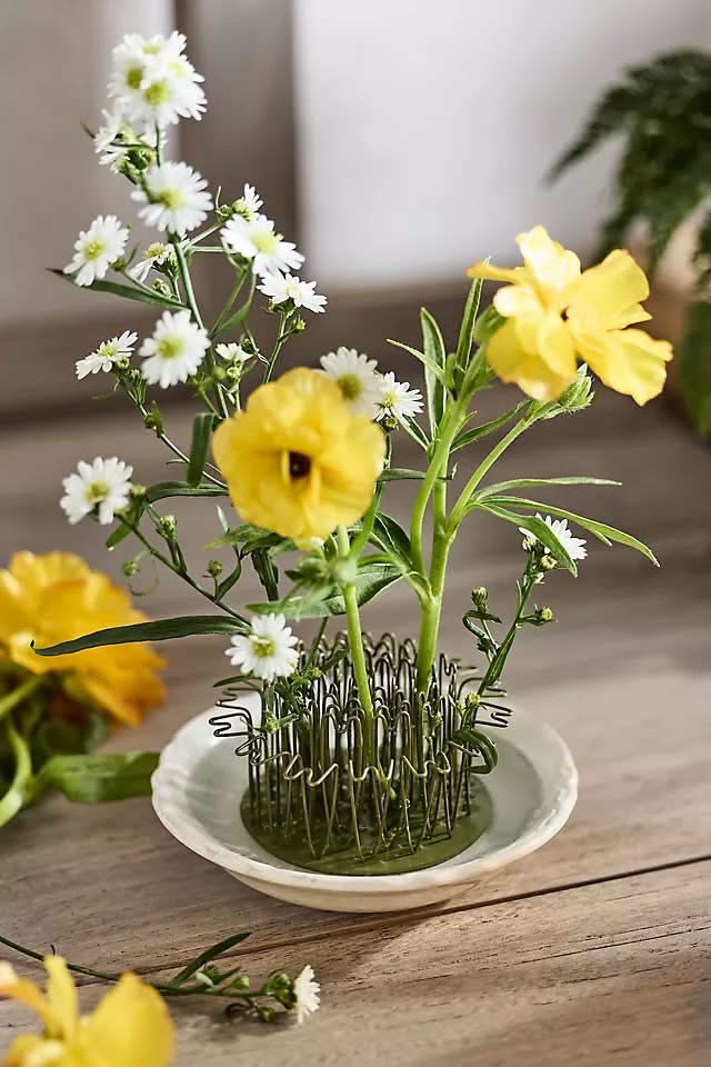 Yellow flowers and white daisies arranged in a wire flower frog on a saucer