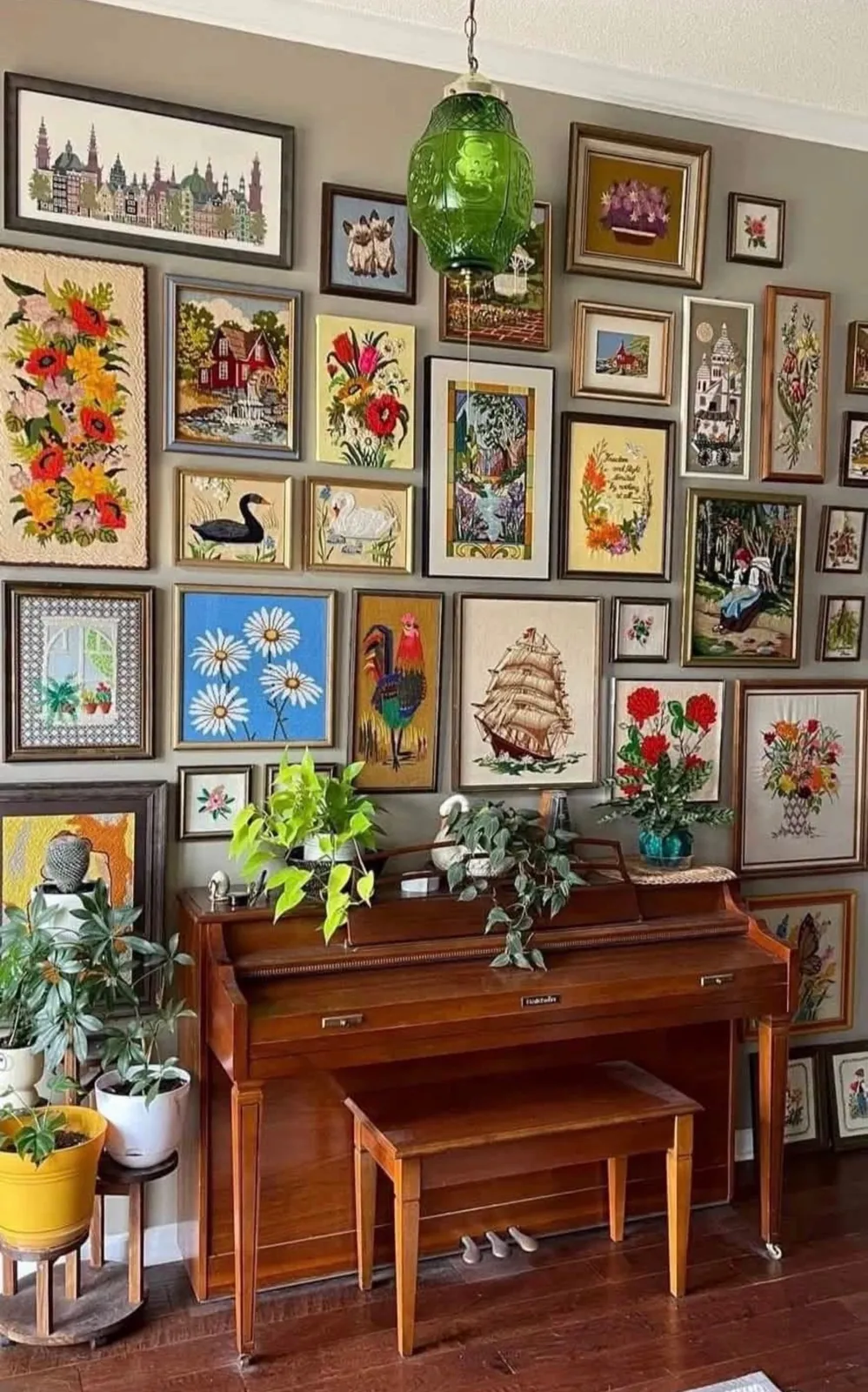 Vintage gallery wall of framed floral, bird and landscape artwork above a wooden piano, with houseplants and a green glass pendant light