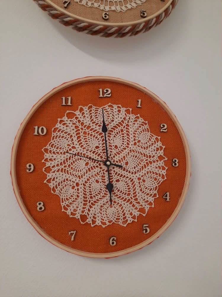 A white doily clock on orange fabric in a wooden hoop