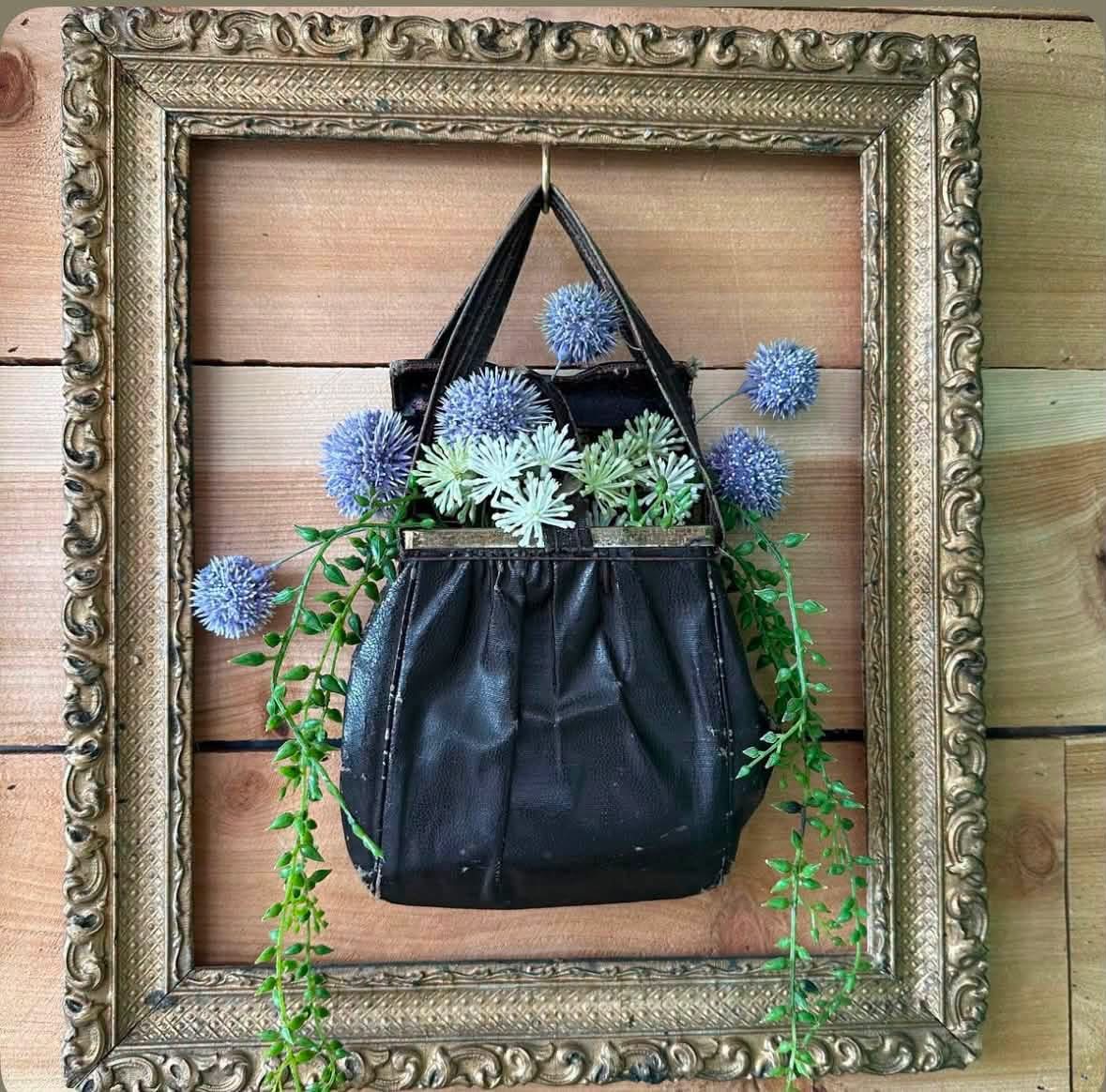 Blue flowers and trailing greenery in a black purse inside a gold frame