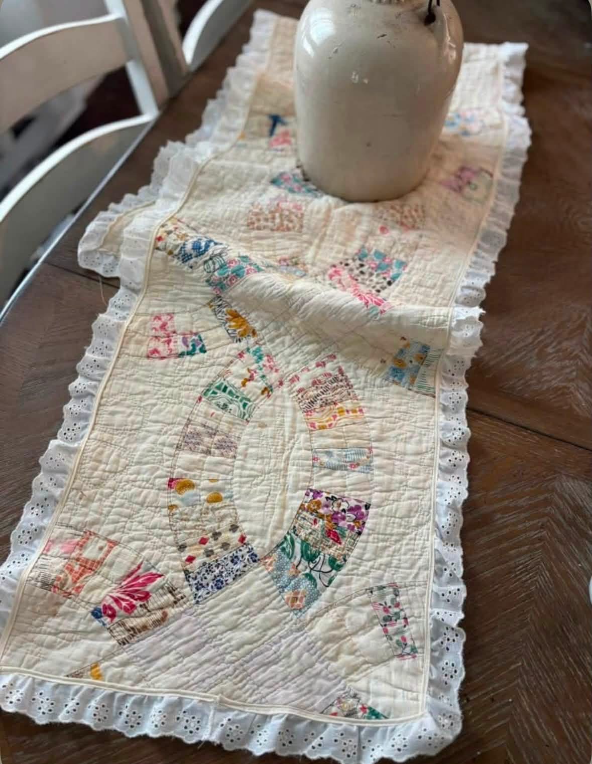 Vintage patchwork quilt table runner with lace trim beneath a cream pitcher